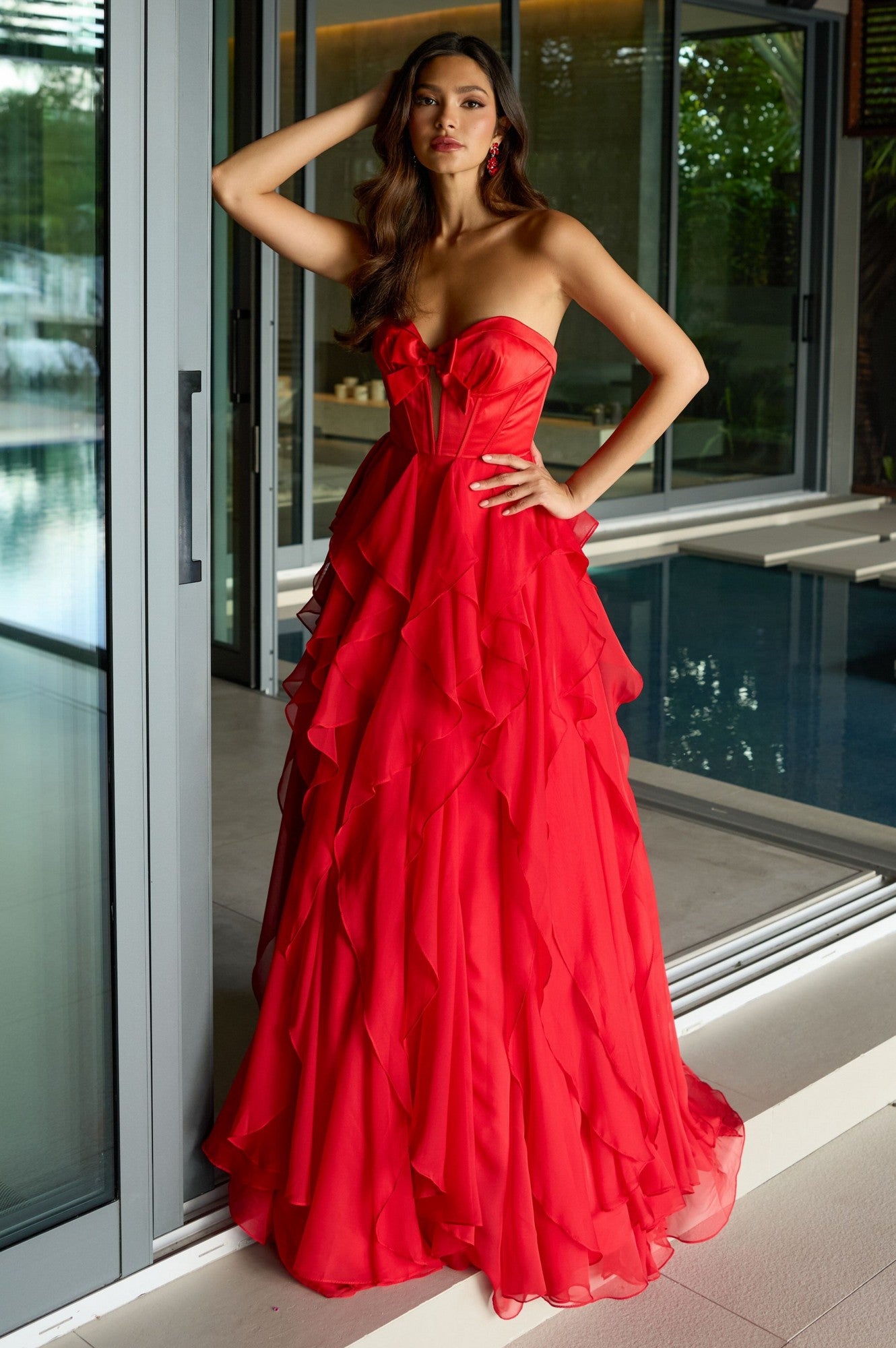 A woman in the red Ava Presley 42209 strapless ruffled prom gown stands by glass doors near an indoor pool, striking a pose with one hand on her head and the other on her hip.